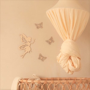 Pu&ograve; includere: Decorazione murale per cameretta beige chiaro con una silhouette di fata in legno e tre ritagli di farfalle in legno. Un baldacchino color crema &egrave; appeso al soffitto e una culla in vimini &egrave; visibile.