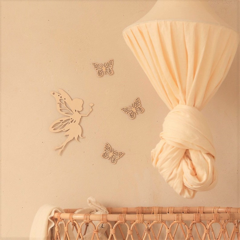 Pu&ograve; includere: Decorazione murale per cameretta beige chiaro con una silhouette di fata in legno e tre ritagli di farfalle in legno. Un baldacchino color crema &egrave; appeso al soffitto e una culla in vimini &egrave; visibile.