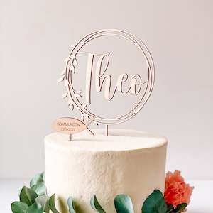 May include: Wooden cake topper with a circle design and the name "Theo" in cursive script. The topper also includes a small fish with the text "Communion 23.04.2022".