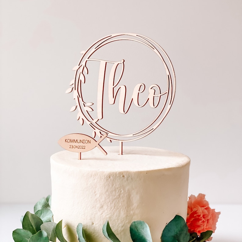 May include: Wooden cake topper with a circle design and the name "Theo" in cursive script. The topper also includes a small fish with the text "Communion 23.04.2022".