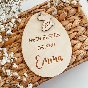 May include: A wooden Easter egg ornament with the text "Mein erstes Ostern Emma" and a small bunny cutout with the year 2022.