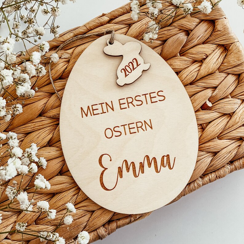 May include: A wooden Easter egg ornament with the text "Mein erstes Ostern Emma" and a small bunny cutout with the year 2022.