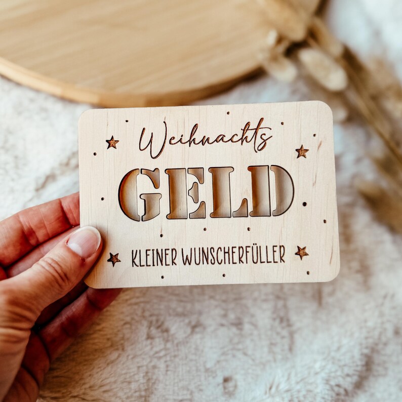 May include: A rectangular wooden plaque with the words "Weihnachts GELD Kleiner Wunscherf&uuml;ller" cut out. The plaque has star and dot accents. The plaque is held by a person.