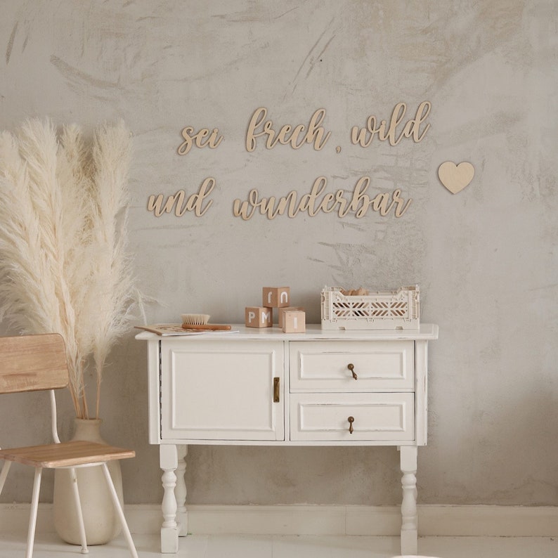 Op de afbeelding: Houten wanddecoratie met de tekst "sei frech. wild und wunderbar" in cursief schrift. De decoratie is getoond op een lichtgrijze muur met een witte commode en een vaas met gedroogd pampasgras.