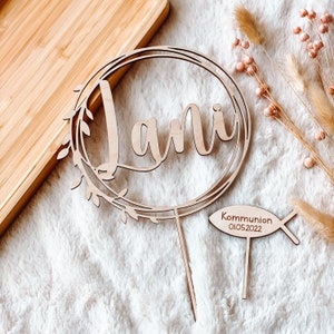 May include: Wooden cake topper with the name "Jani" in a cursive font, surrounded by a wreath of leaves. A separate wooden fish-shaped topper reads "Communion 01.05.2022".