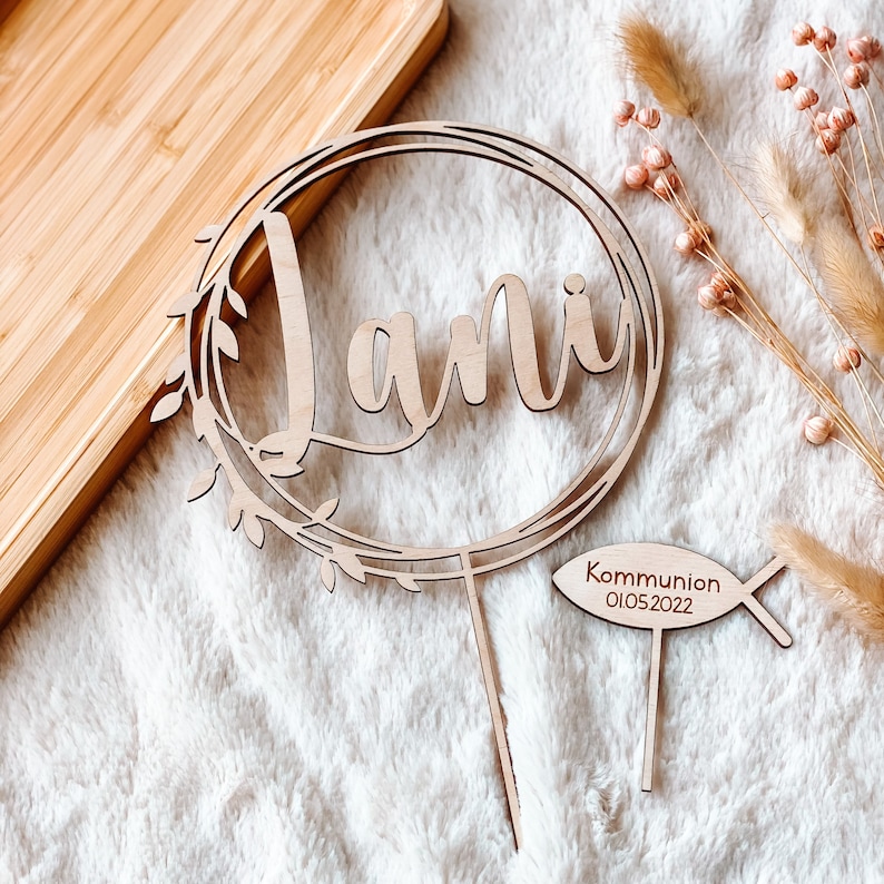May include: Wooden cake topper with the name "Jani" in a cursive font, surrounded by a wreath of leaves. A separate wooden fish-shaped topper reads "Communion 01.05.2022".