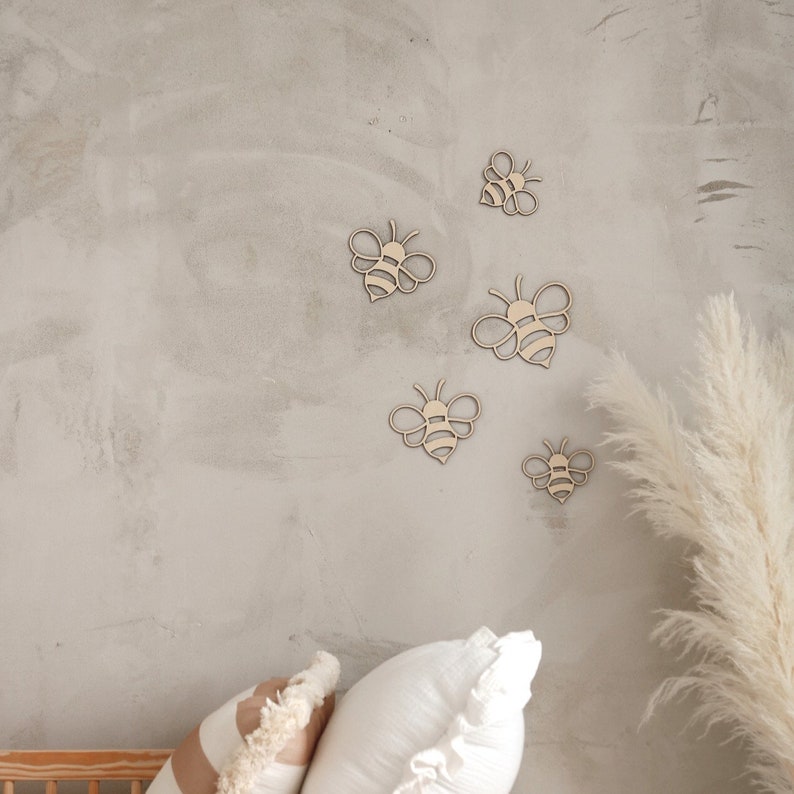 May include: Five wooden bee cutouts in various sizes are arranged on a light gray textured wall. The bees are all facing the same direction and have a simple, minimalist design.