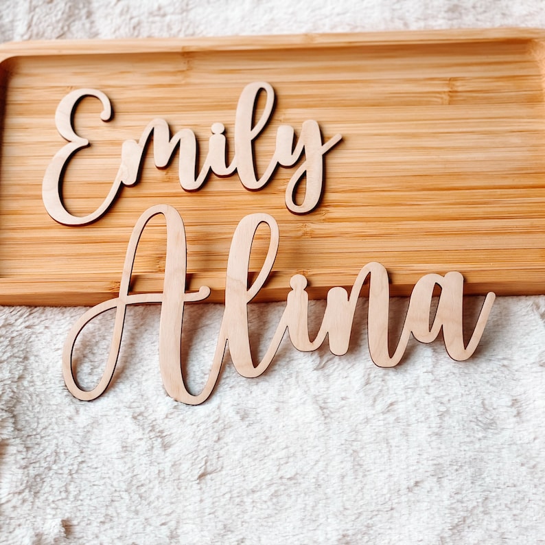 Puede incluir: Dos letreros de madera con los nombres "Emily" y "Alina" se muestran en una bandeja de madera.