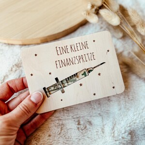 May include: A wooden card with the text "EINE KLEINE FINANZSPRITZE" and a syringe design filled with dollar bills. The card is light-colored with small star accents.