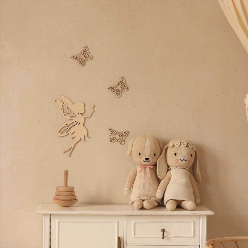 Pu&ograve; includere: Una scena di cameretta con decorazioni murali in legno a forma di farfalle e fatine, un giocattolo impilabile in legno e due bambole di peluche: un cane e un coniglio. Le bambole indossano abiti e il coniglio ha una corona floreale.