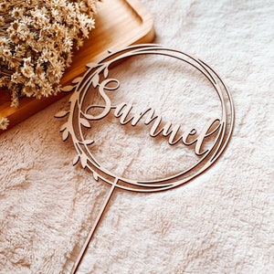 May include: A wooden cake topper with a circular frame and leafy details. The name "Samuel" is written in cursive script inside the frame.
