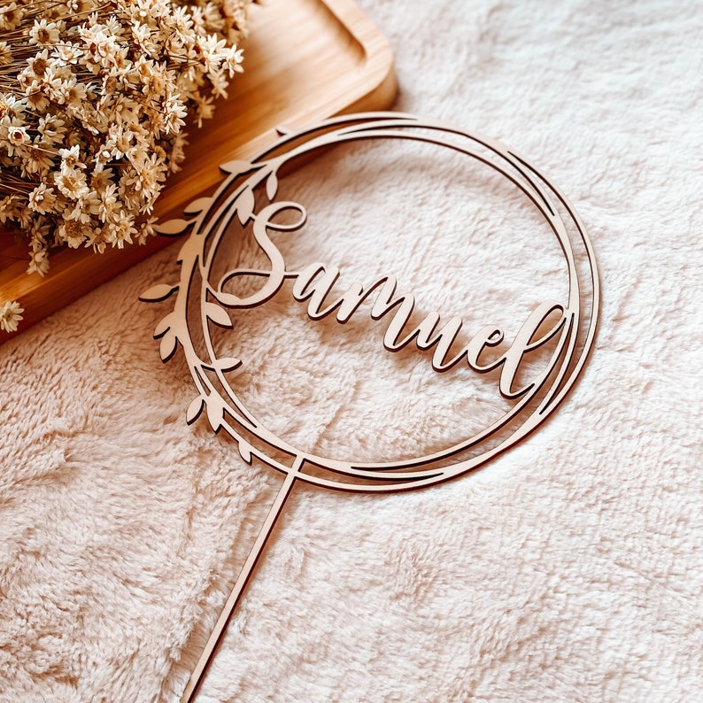 May include: A wooden cake topper with a circular frame and leafy details. The name "Samuel" is written in cursive script inside the frame.