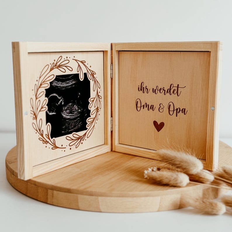 Pu&ograve; includere: Una scatola di legno color chiaro, aperta per rivelare un'immagine ecografica incorniciata da un disegno a foglie. Il lato destro della scatola presenta il testo "ihr werdet Oma & Opa" con un simbolo a cuore. La scatola &egrave; su una superficie di legno.