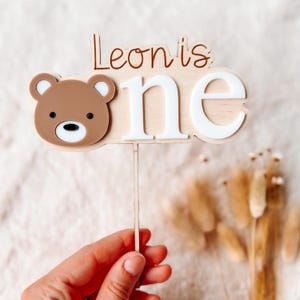 Puede incluir: Adorno de pastel de madera con la cara de un oso marr&oacute;n y las palabras "Leon is one" en marr&oacute;n y blanco. El oso tiene un hocico blanco y ojos negros. El adorno se sujeta con un palito de madera.