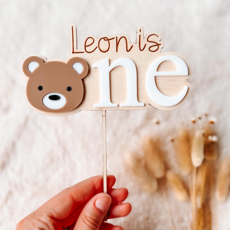 Puede incluir: Adorno de pastel de madera con la cara de un oso marr&oacute;n y las palabras "Leon is one" en marr&oacute;n y blanco. El oso tiene un hocico blanco y ojos negros. El adorno se sujeta con un palito de madera.