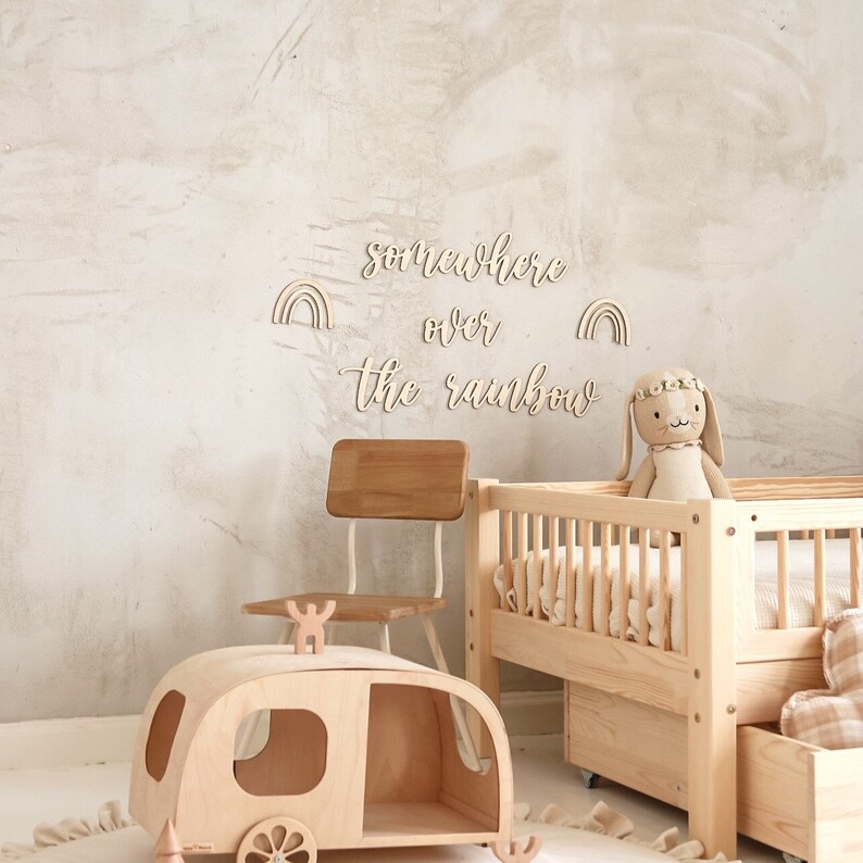 K&ouml;nnte beinhalten: Ein Spielzimmer aus Holz mit einem Holzbettchen, einem Holzstuhl, einem Holzspielzeug-Wohnwagen und einem Holzhasenspielzeug. An der Wand befindet sich ein Holzschild mit der Aufschrift "somewhere over the rainbow" und zwei Regenbogen-Dekorationen.