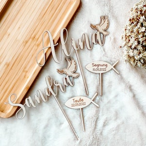 May include: Three wooden cake toppers featuring the names Alina, Sandra, and a dove with an olive branch. The toppers also include a wooden fish with the words "Segnung 15.05.2022" and "Taufe 22.05.2022".