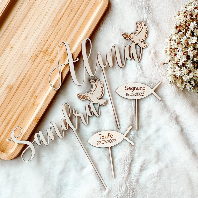 May include: Three wooden cake toppers featuring the names Alina, Sandra, and a dove with an olive branch. The toppers also include a wooden fish with the words "Segnung 15.05.2022" and "Taufe 22.05.2022".
