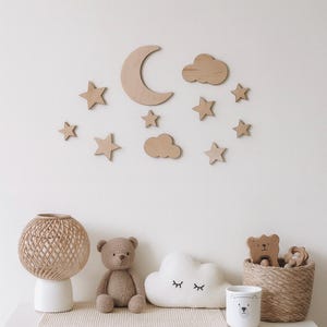 May include: A nursery wall decor set featuring wooden stars, clouds, and a crescent moon. Below, a teddy bear, cloud-shaped pillow, and woven basket with wooden toys sit on a white surface. A woven lamp and a bear-themed mug are also present.