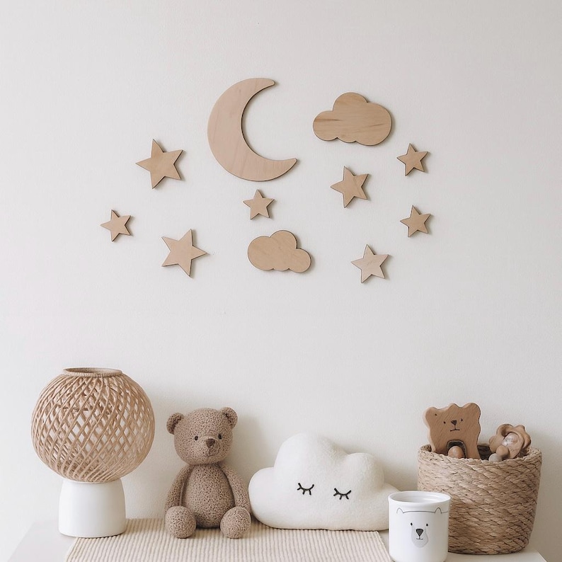 May include: A nursery wall decor set featuring wooden stars, clouds, and a crescent moon. Below, a teddy bear, cloud-shaped pillow, and woven basket with wooden toys sit on a white surface. A woven lamp and a bear-themed mug are also present.