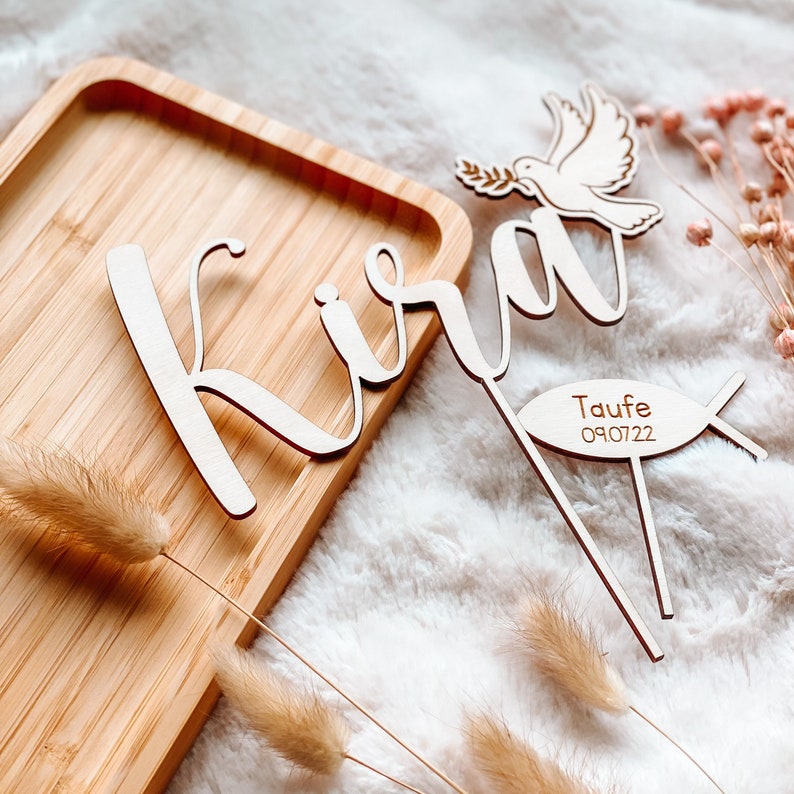 May include: Wooden cake topper with the name "Kira" in cursive script, a dove with an olive branch, and a fish with the text "Taufe 09.07.22".