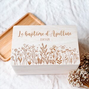 May include: A light-colored wooden box with a floral design and the text "Le bapt&ecirc;me d'Apolline 19.09.21" engraved on the lid. The box has a rectangular shape and a natural wood grain finish.