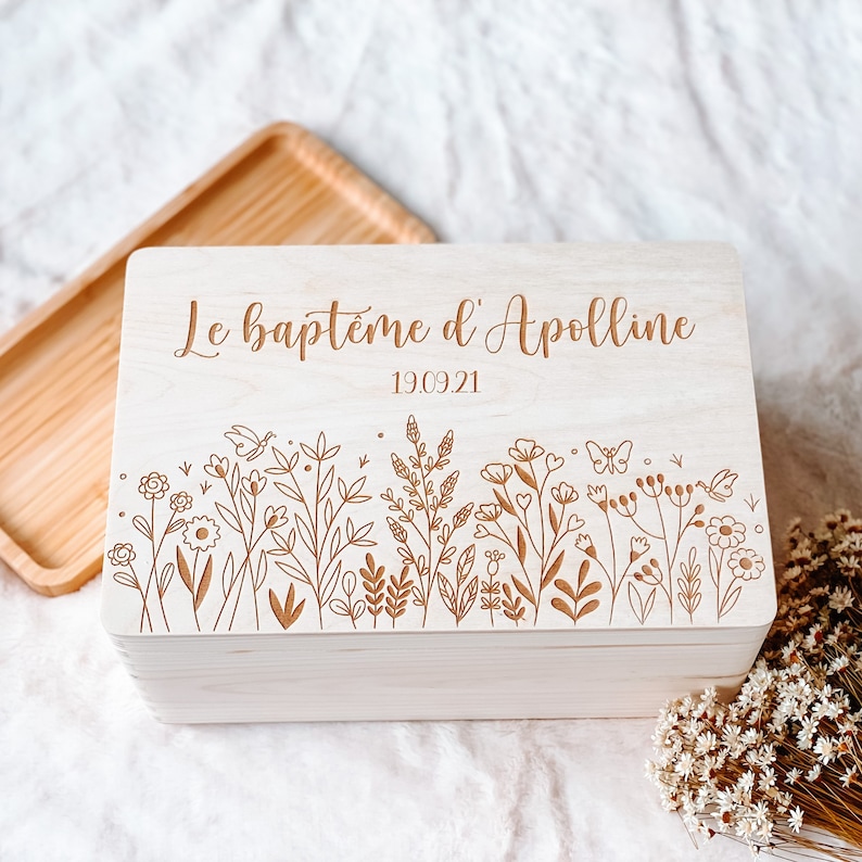 May include: A light-colored wooden box with a floral design and the text "Le bapt&ecirc;me d'Apolline 19.09.21" engraved on the lid. The box has a rectangular shape and a natural wood grain finish.
