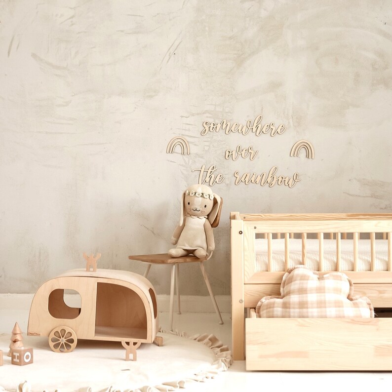 K&ouml;nnte beinhalten: Ein h&ouml;lzerner Spielzeug-Wohnwagen mit einer Hasenpuppe, die auf einem Holzstuhl sitzt. Die Wand hinter den Spielzeugen hat ein Holzschild mit der Aufschrift "somewhere over the rainbow" und zwei Regenbogen-Ausschnitten auf beiden Seiten des Schildes.