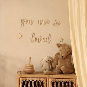 May include: Wooden wall decor with the words "you are so loved" in cursive script. The decor is displayed on a beige wall with two wooden stars and a wicker shelf with a wooden toy and two stuffed animals.