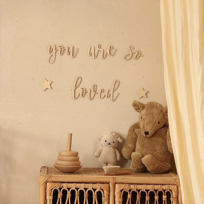 May include: Wooden wall decor with the words "you are so loved" in cursive script. The decor is displayed on a beige wall with two wooden stars and a wicker shelf with a wooden toy and two stuffed animals.