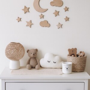 May include: A nursery scene with wooden moon, stars, and cloud wall decor. A white dresser holds a woven lamp, teddy bear, cloud pillow, bear-themed cup, and a woven basket with wooden bear toys. Soft, neutral tones create a calming atmosphere.