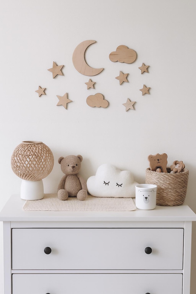 May include: A nursery scene with wooden moon, stars, and cloud wall decor. A white dresser holds a woven lamp, teddy bear, cloud pillow, bear-themed cup, and a woven basket with wooden bear toys. Soft, neutral tones create a calming atmosphere.