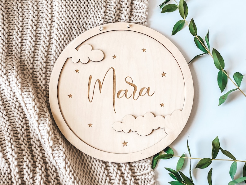 May include: A round wooden sign with the name "Mara" written in cursive script. The sign is decorated with clouds and stars, and has a hole at the top for hanging.