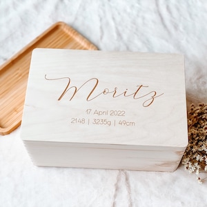 May include: A light-colored wooden box with the name "Moritz" engraved on the lid. The date "17. April 2022" and measurements "2148 | 3235g | 49cm" are also engraved. The box is rectangular with a slightly rounded top.