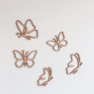 May include: Five wooden butterfly cutouts in various designs and sizes. The butterflies are a light brown color and have a simple, outlined appearance. They are arranged against a plain white background, creating a minimalist aesthetic.