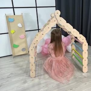 May include: A wooden play structure with a climbing wall and a ladder. A young girl is playing inside the structure. The structure is painted in natural wood tones and has colorful accents.