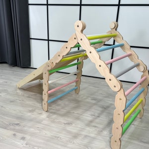Könnte beinhalten: Eine Holz-Kletterstruktur mit Rampe und Kletterbogen. Der Bogen hat farbige Sprossen in Pastelltönen von Rosa, Blau, Grün und Grau. Die Struktur ist aus hellem Holz gefertigt und für Kinder konzipiert.