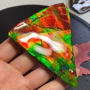 May include: A triangular piece of iridescent opal with red, green, and blue hues. The opal has a smooth, polished surface and is a vibrant, colorful gemstone.