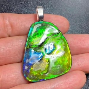 Puede incluir: Colgante de ammolita iridiscente con una fianza de plata. La piedra preciosa muestra un espectro de colores, incluyendo verde, azul y dorado. La superficie pulida revela patrones y texturas naturales, creando una joya única y llamativa.