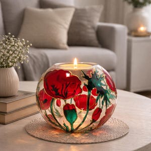 May include: A spherical glass candle holder featuring hand-painted red poppy flowers and green foliage with gold outlines. The clear glass vessel contains a lit candle, casting a warm glow through the floral artwork on the rounded surface.