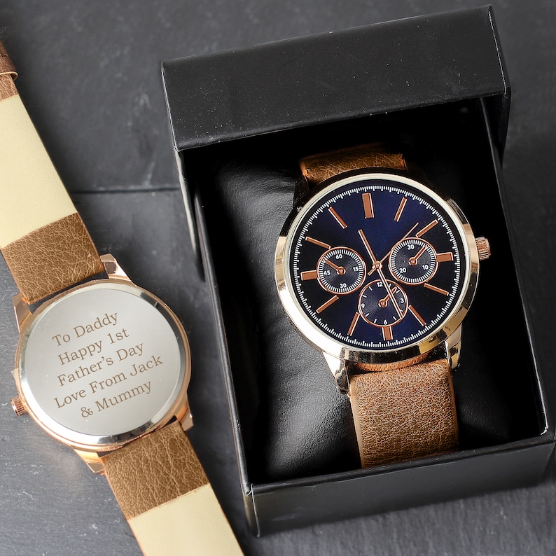 May include: A brown leather wristwatch with a gold-tone case and blue face. The watch has a personalized message on the back that reads "To Daddy Happy 1st Father's Day Love From Jack & Mummy".