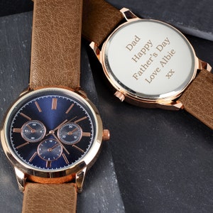 May include: A gold-tone watch with a brown leather band. The watch face is blue with three smaller dials. The back of the watch has a personalized message that reads "Dad Happy Father's Day Love Albie XX".