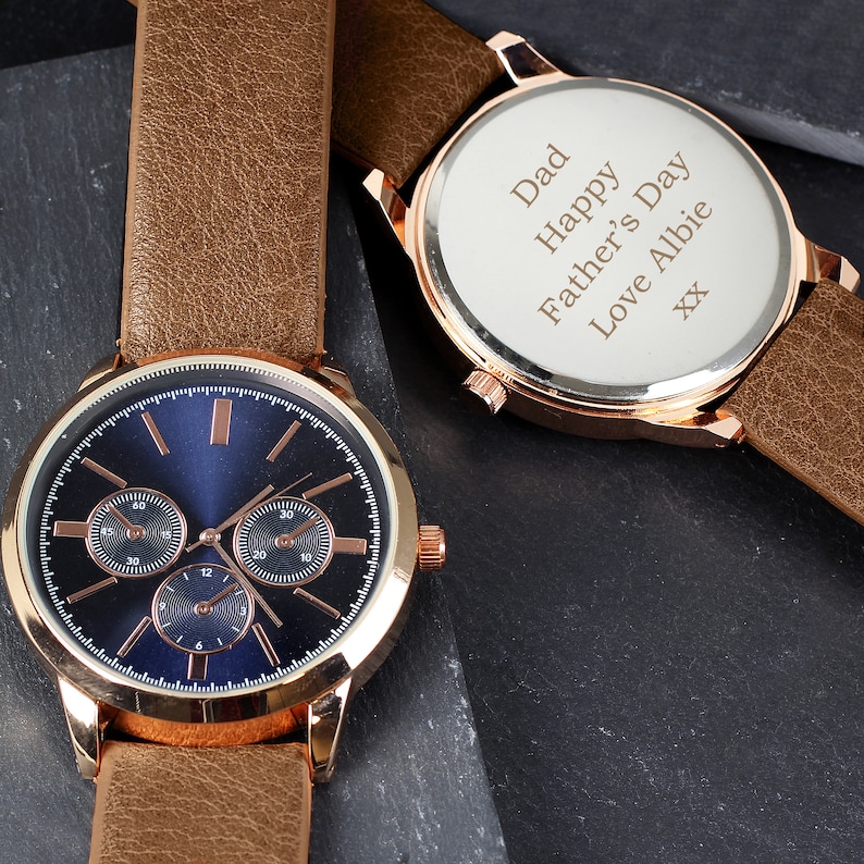 May include: A gold-tone watch with a brown leather band. The watch face is blue with three smaller dials. The back of the watch has a personalized message that reads "Dad Happy Father's Day Love Albie XX".