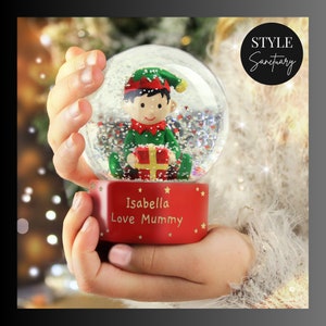 May include: A red snow globe with a small elf figurine holding a gift inside. The base of the snow globe is decorated with gold stars and the text "Isabella Love Mummy".