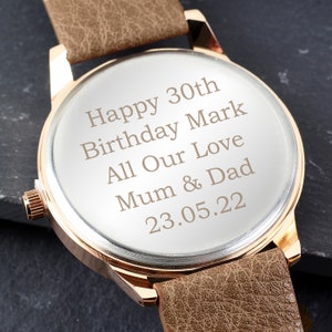 May include: A brown leather wristwatch with a gold-tone case. The watch face is engraved with the message "Happy 30th Birthday Mark All Our Love Mum & Dad 23.05.22".
