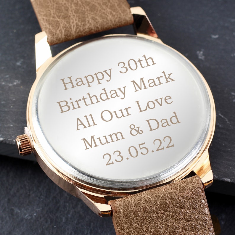 May include: A brown leather wristwatch with a gold-tone case. The watch face is engraved with the message "Happy 30th Birthday Mark All Our Love Mum & Dad 23.05.22".