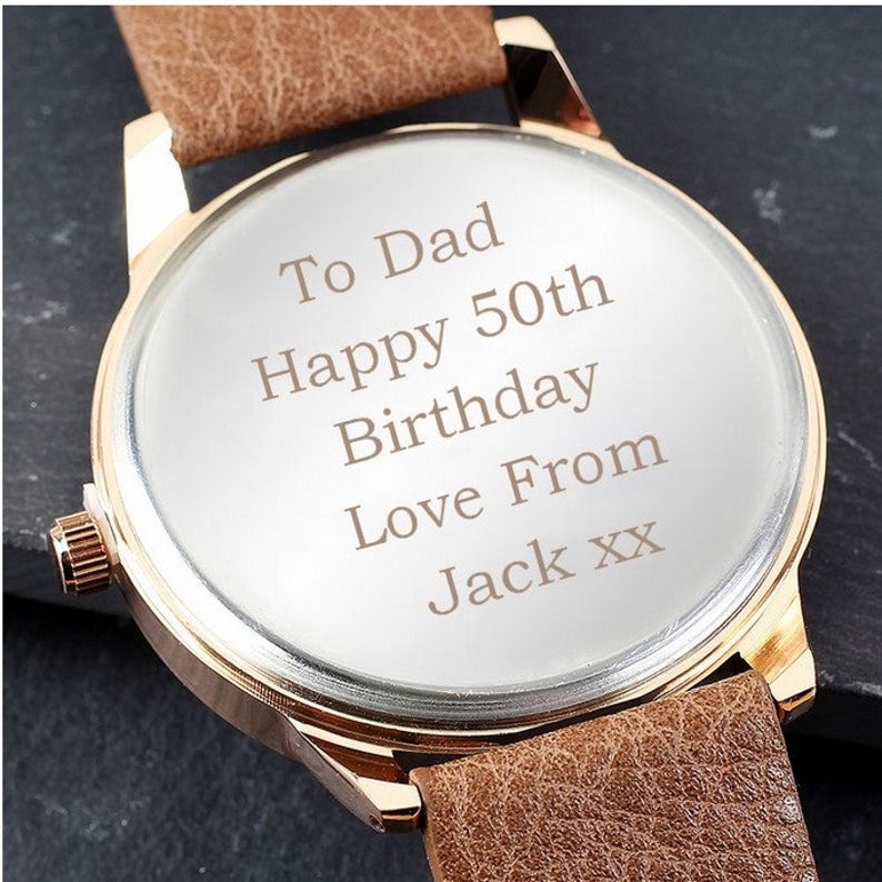 May include: A gold-tone watch with a brown leather band. The watch face is engraved with the message "To Dad Happy 50th Birthday Love From Jack xx".