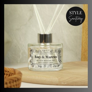 May include: A clear glass diffuser bottle with a silver cap and black reeds. The bottle has a white label with black text that reads "Congratulations Amy & Marcus On Your Wedding Day 14th August 2024". The label is surrounded by a floral design.