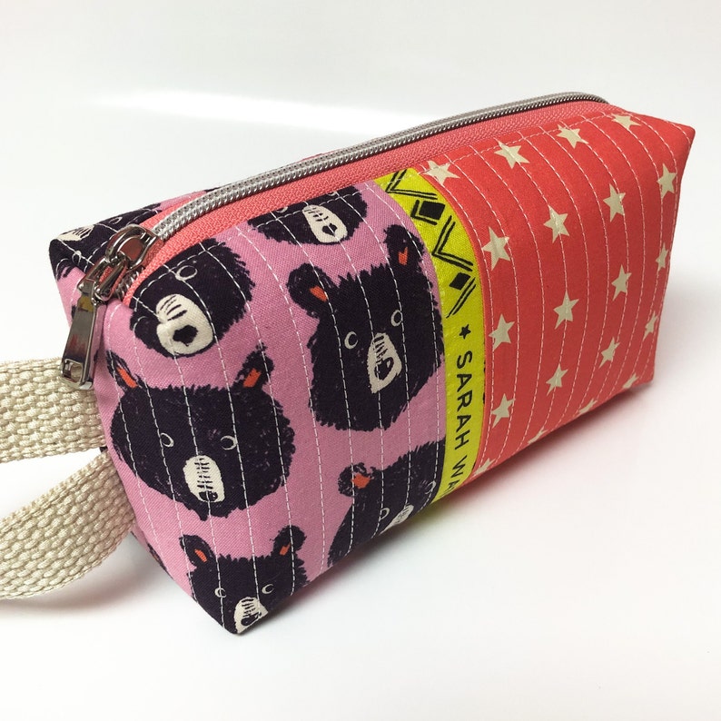 May include: Pink zippered pouch with a black bear print and a yellow and red stripe with the text "SARAH W" on the side. The pouch has a beige strap.
