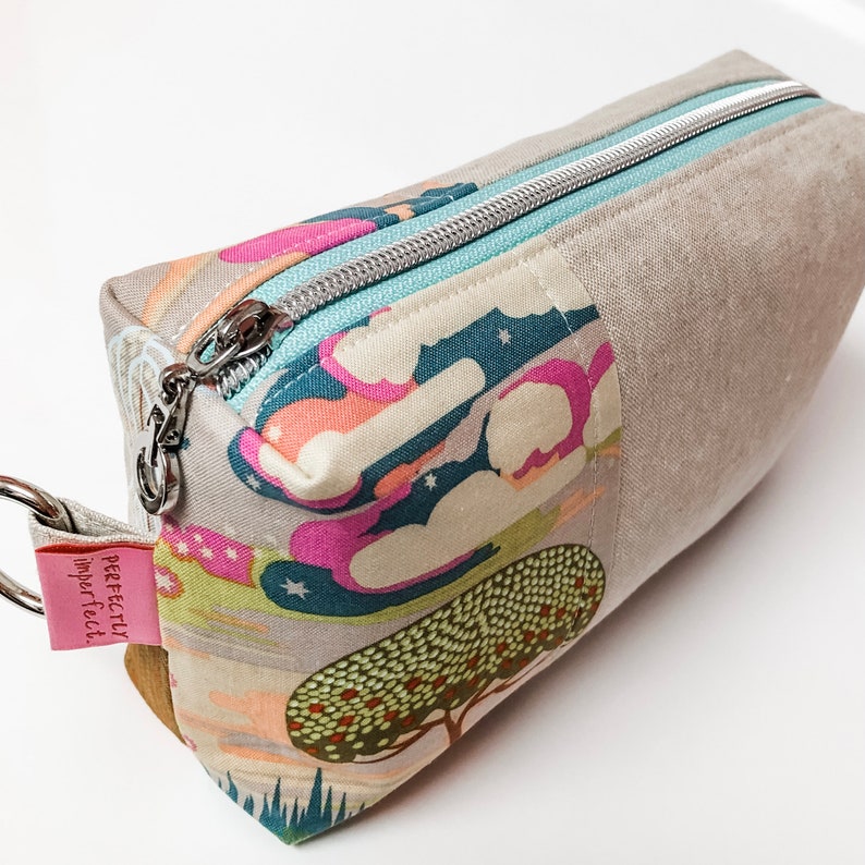 May include: A beige zippered pouch with a colorful fabric lining featuring a whimsical design of clouds, stars, and trees. The pouch has a silver zipper and a brown leather tag with the text "perfectly imperfect".